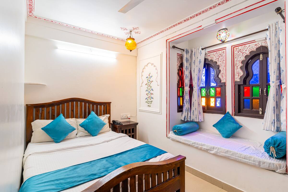 Un dormitorio con cama de madera, una mesita de noche, cortinas decorativas en la ventana y una lámpara fijada a la pared.