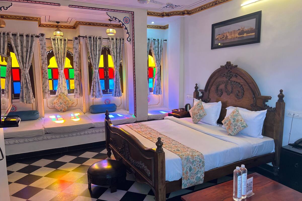 Una habitación de hotel con piso a cuadros, una cama de madera con ropa de cama floral, una mesita de noche con botellas de agua y un cuadro colgado en la pared.