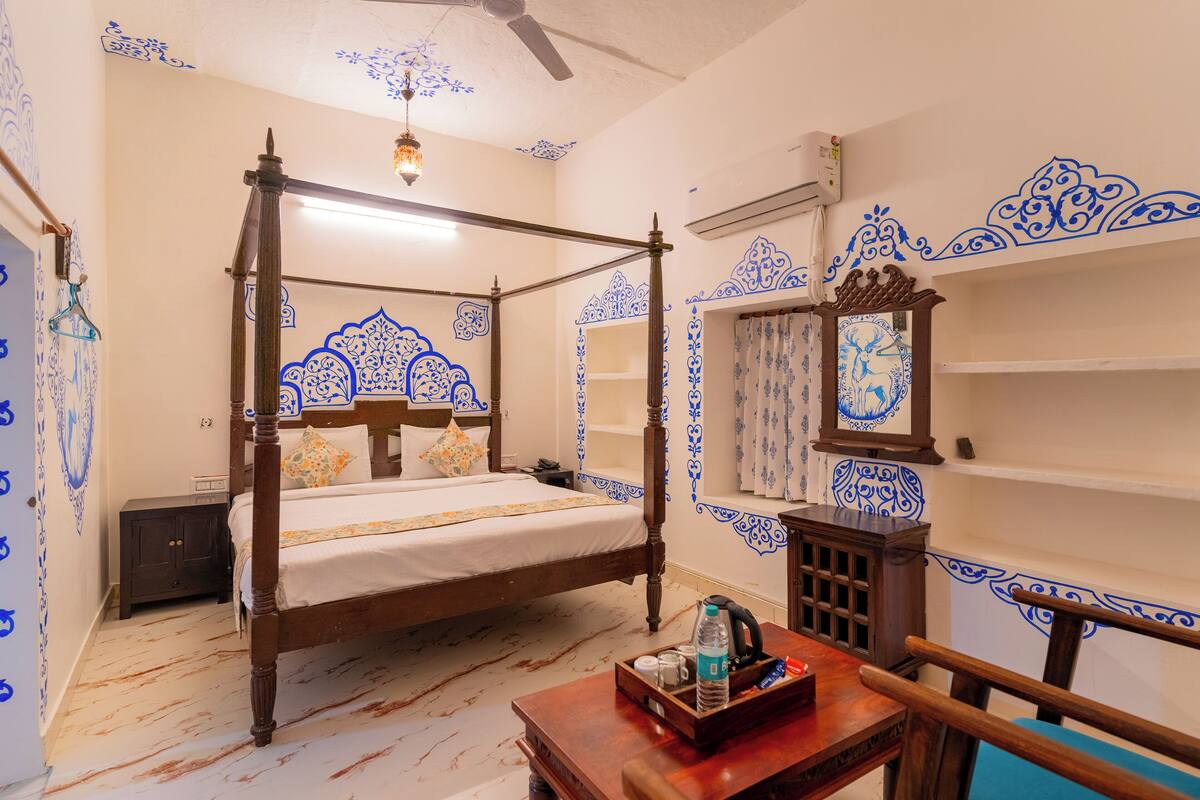 Una cama con dosel de madera y colcha blanca, una mesita de noche de madera con una lámpara, una pequeña mesa con una bandeja, y patrones azules decorativos en las paredes y el techo.