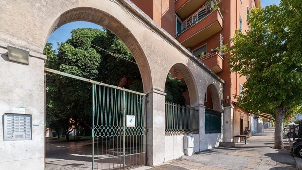 A street view with a gated entrance, a building with arched windows, and a tree-lined sidewalk.