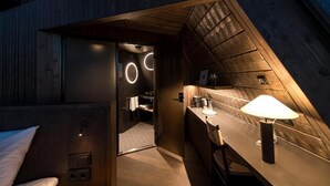 Una habitación moderna, con poca luz, techo de madera, cama, mesita con lámpara y baño visible a través de una puerta abierta.
