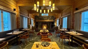 Un comedor acogedor con paneles de madera, suelo de piedra, una chimenea central y varias mesas dispuestas para comer.