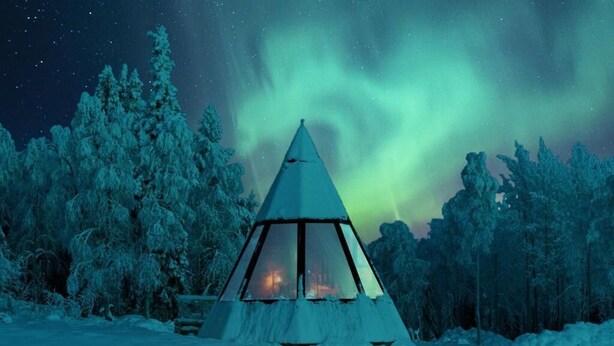 Tienda triangular cubierta de nieve con fuego en su interior, rodeada por un bosque bajo un cielo estrellado.