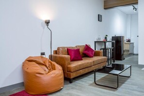 Una sala moderna con un sofá de cuero, un sillón de peluche, una mesa de centro de vidrio y una cocina al fondo.