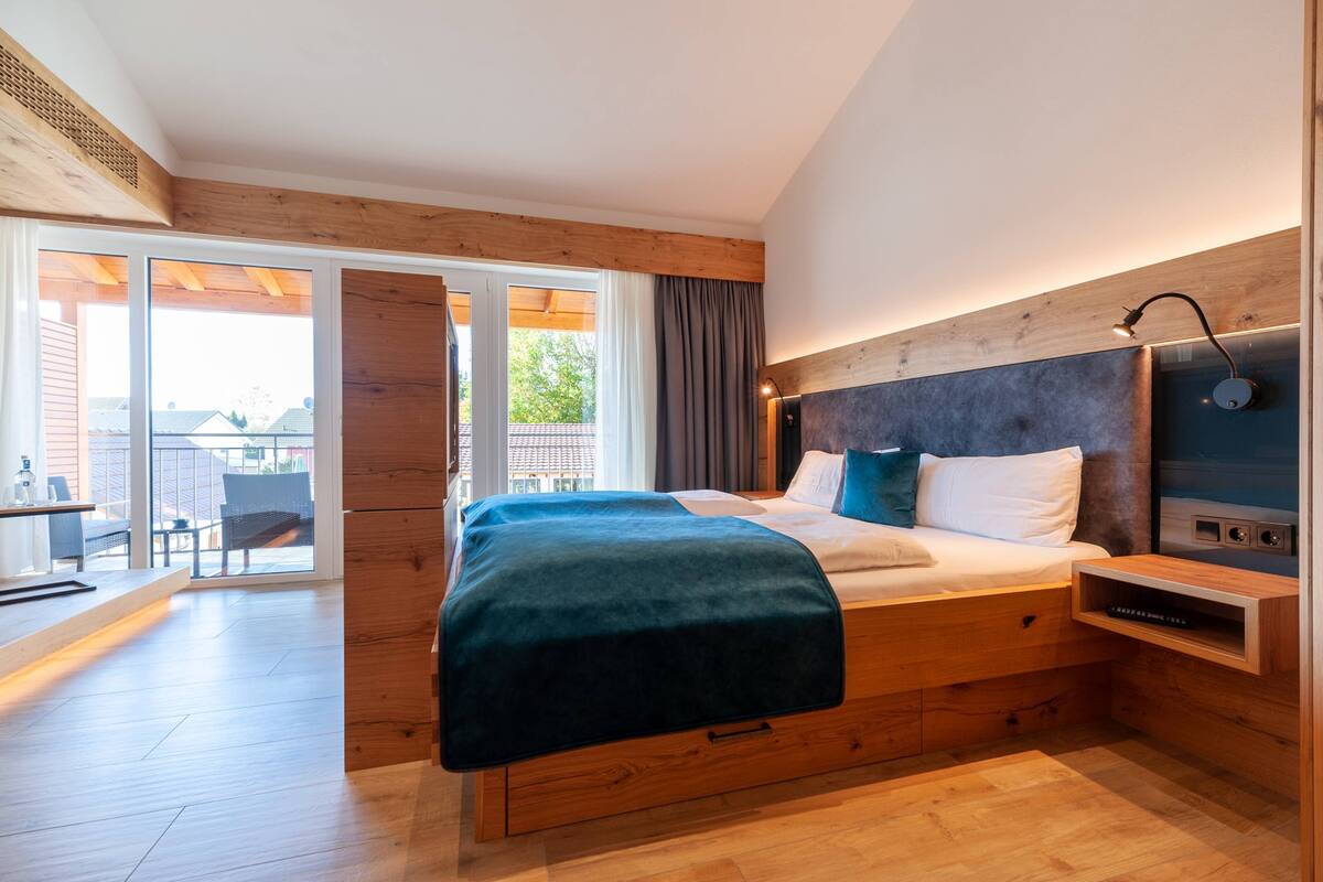 Un dormitorio moderno con cabecera de madera, una cama grande con manta azul, una mesita de noche y vistas al exterior a través de una puerta corrediza de vidrio.