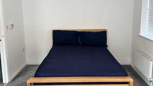 Un lit simple avec un cadre en bois et une literie bleu foncé, dans une pièce aux murs blancs et dotée d’une fenêtre avec des stores.