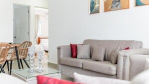 Una sala de estar con un sofá gris, cojines rojos, una mesa de centro de cristal, sillas de mimbre y una puerta al fondo.
