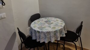 A small dining area with a round table covered by a floral tablecloth, two black chairs, a white cabinet above the table, and a wall-mounted telephone.