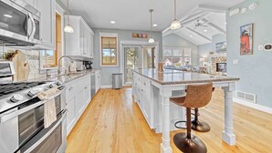 A modern kitchen with white cabinetry, a large island with bar stools, a stainless steel stove, and a view into the dining area. - Harbor Views, Hot Tub, Walk to Beach, Dog Friendly (South Haven)