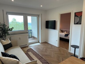 Ein Zimmer mit Sofa, Fernseher, Bett und Blick durch eine Schiebetür.