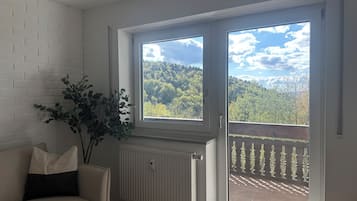A room with a white brick wall, a radiator, a beige sofa with a black cushion, a potted plant, and a sliding glass door leading to a balcony with a view of a forested hill.