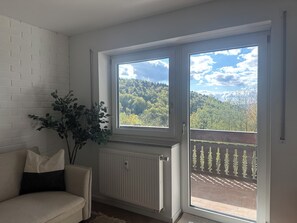 Ein Zimmer mit weißer Ziegelwand, einem Heizkörper, einem beigefarbenen Sofa mit schwarzem Kissen, einer Topfpflanze und einer Glasschiebetür, die auf einen Balkon mit Blick auf einen bewaldeten Hügel führt.