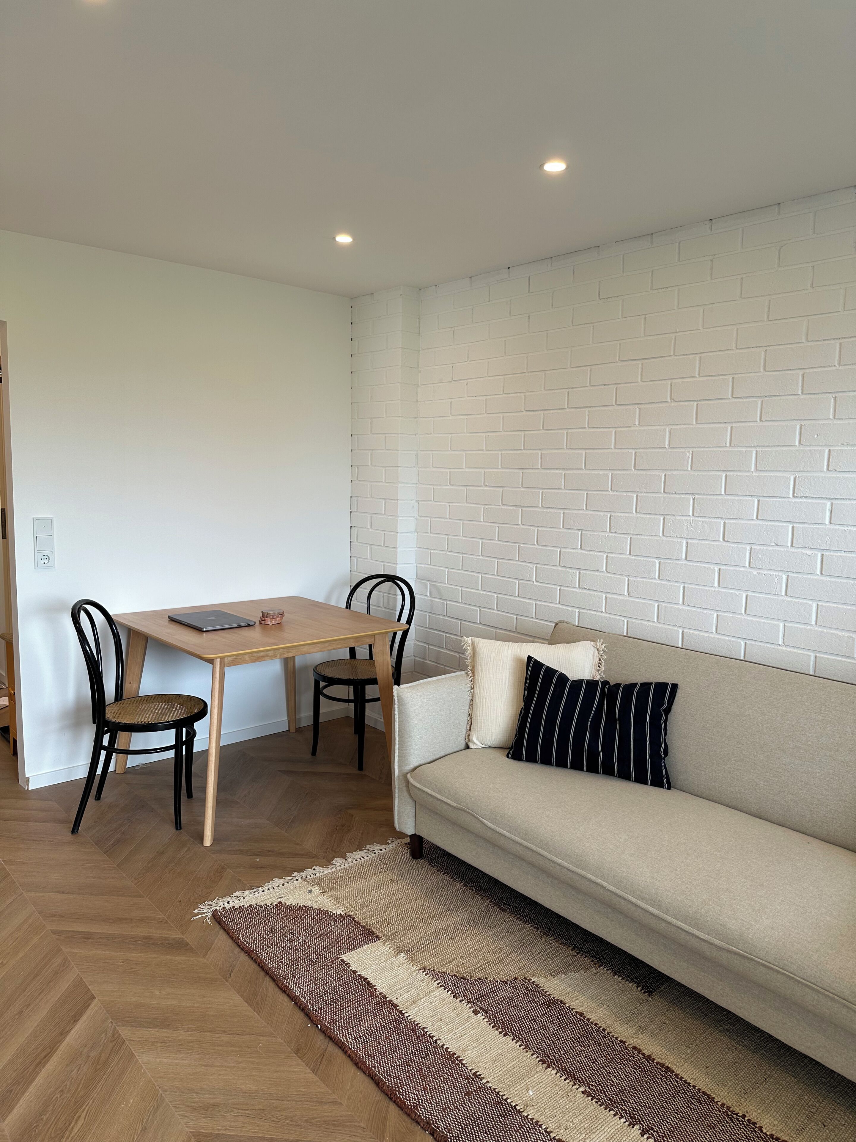 Ein Zimmer mit einer Couch, einem Esstisch mit Stühlen, einer weißen Ziegelwand und einem gemusterten Teppich.