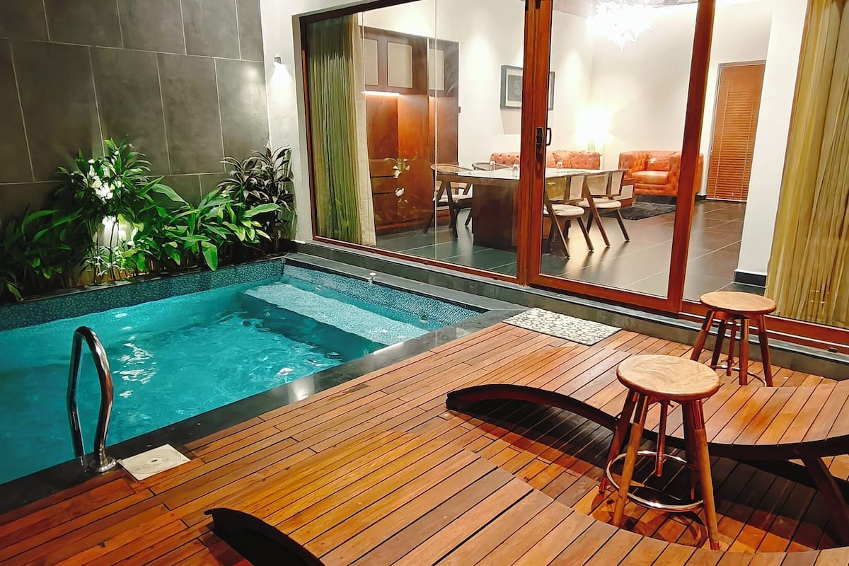 A swimming pool with a wooden deck, a bar area, and a dining area visible through a glass door.