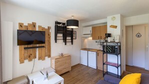 Un espace de vie compact avec un téléviseur fixé sur un mur recouvert de panneaux en bois, un petit meuble en bois, une table à manger avec des chaises, un coin cuisine équipé d’une machine à café et d’un four à micro-ondes, et un coussin jaune sur un canapé.