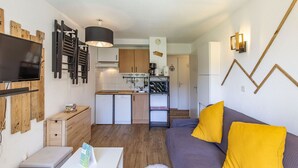 Un espace de vie compact comprenant un coin cuisine, un canapé avec des coussins jaunes, un téléviseur et une décoration murale en bois.