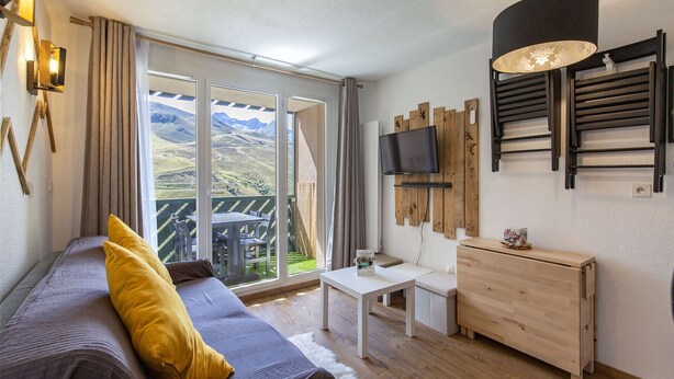 Une pièce confortable avec un canapé, une petite table, un téléviseur fixé sur un panneau en bois et un balcon offrant une vue sur les montagnes.