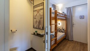 Une petite pièce avec un lit superposé, une porte dotée d’un code QR, un porte-rouleau de papier toilette et un tableau encadré au mur.
