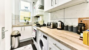 A modern kitchen with white cabinets, a wooden countertop, a washing machine, a coffee maker, and a kettle. - Drayton Green Road (London)