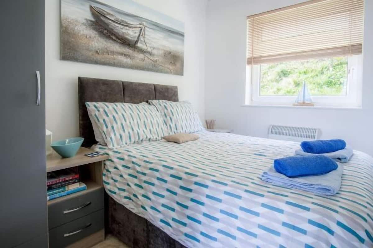 A bedroom with a bed, a window with blinds, a bedside table with a bowl, and a bookshelf.