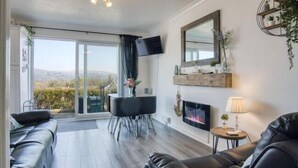 Een moderne woonkamer met een open haard, een flatscreen televisie en een eethoek met een glazen deur naar een balkon.
