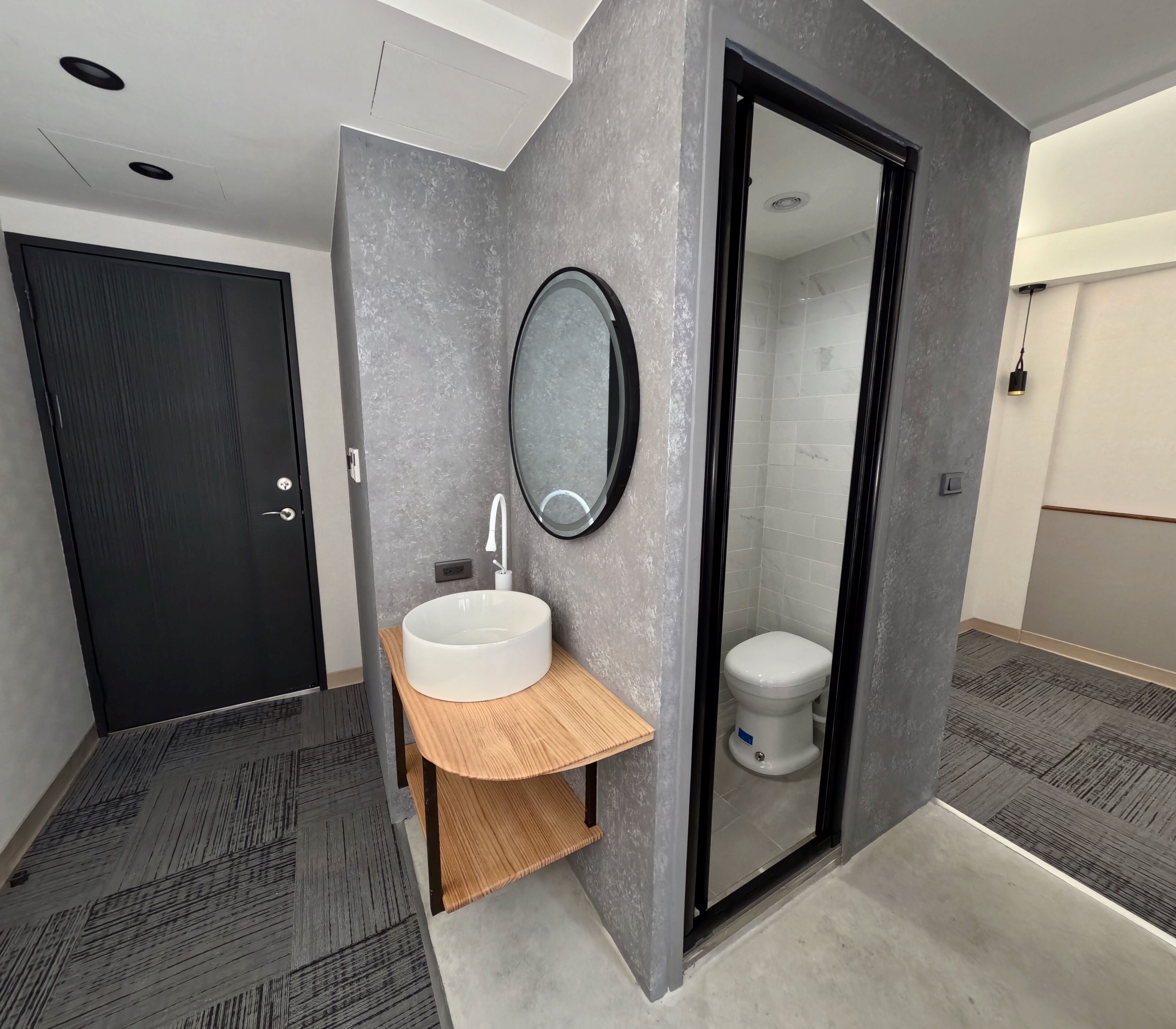 A modern bathroom with a grey textured wall, a round mirror, a white sink with a wooden shelf, and a toilet visible through a glass door.