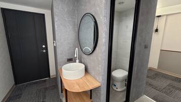 A modern bathroom with a grey textured wall, a round mirror, a white sink with a wooden shelf, and a toilet visible through a glass door.