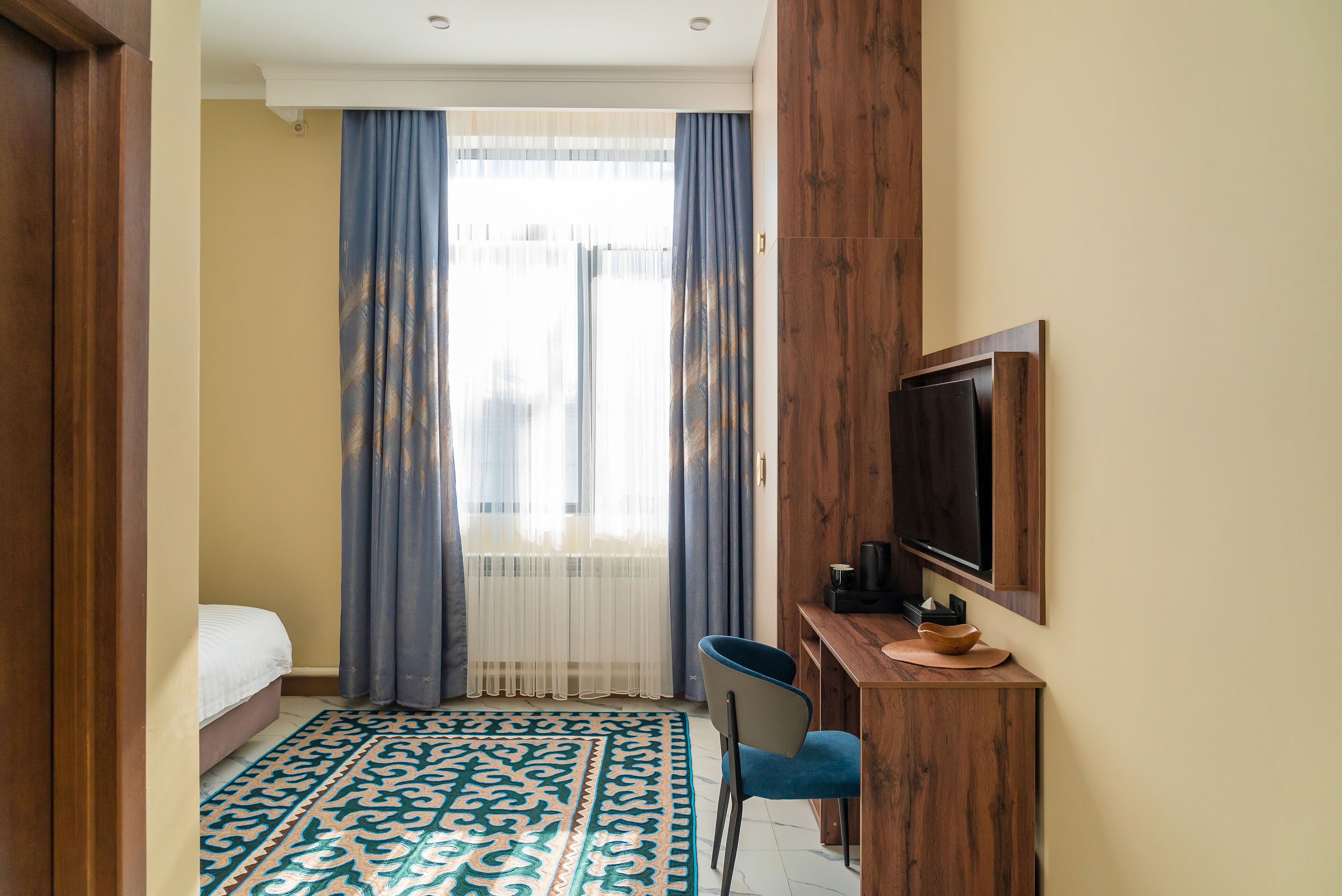 A hotel room with a bed, a desk with a chair, a television, and a patterned rug.