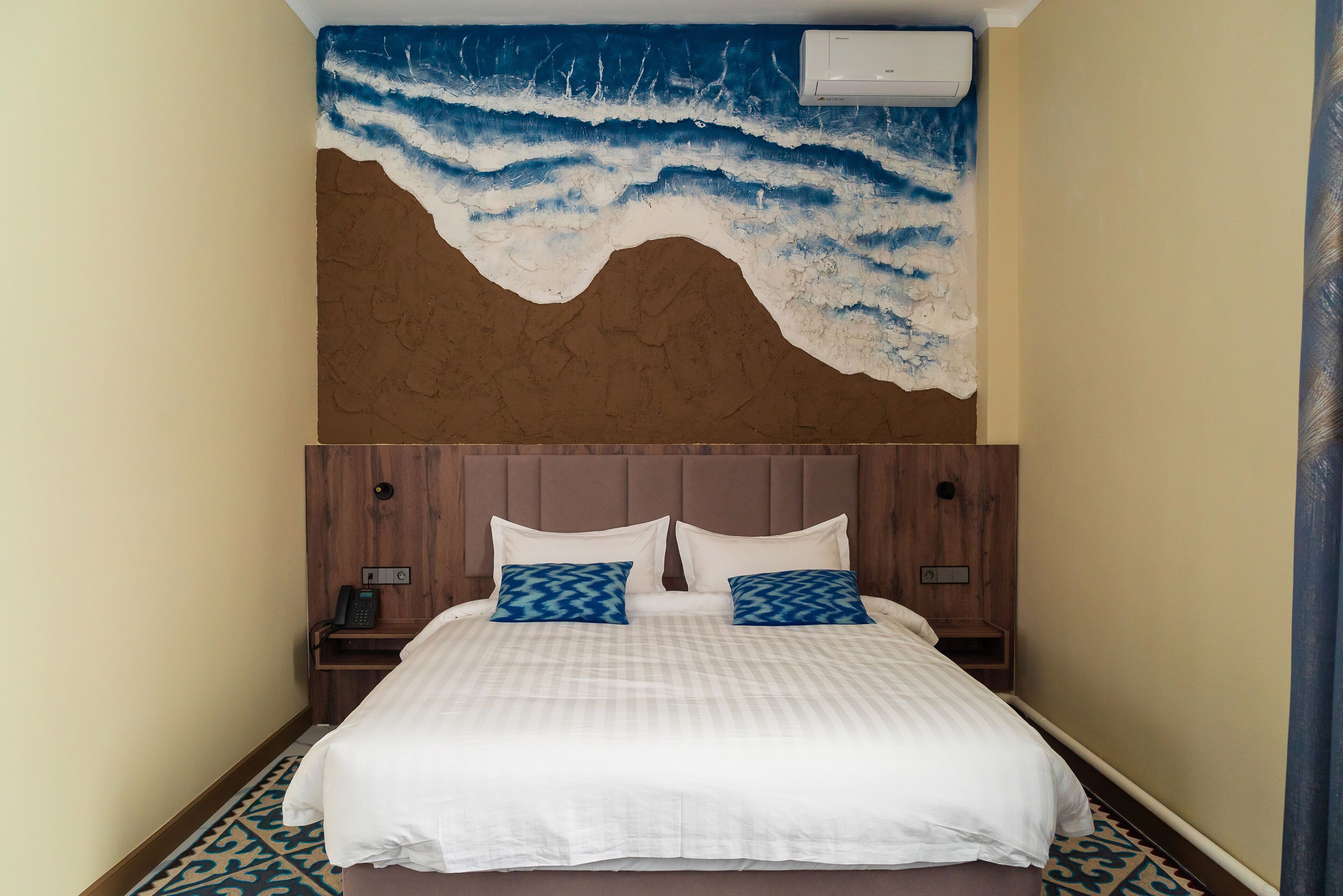 Una habitación de hotel con cama, cabecera, un mural en la pared y una alfombra estampada.