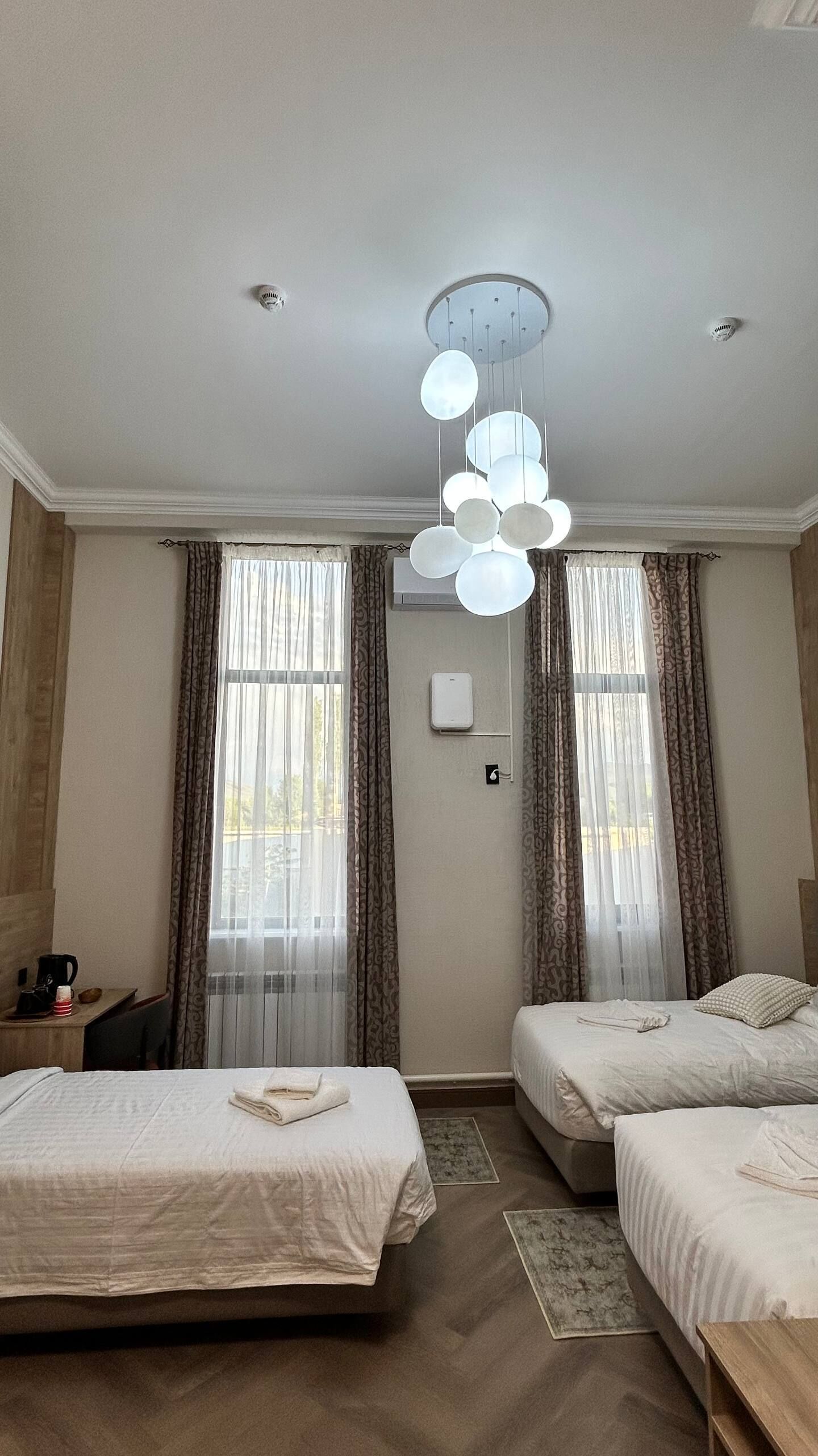 A hotel room with two beds, a modern chandelier, large windows with curtains, and a small table with a coffee maker.