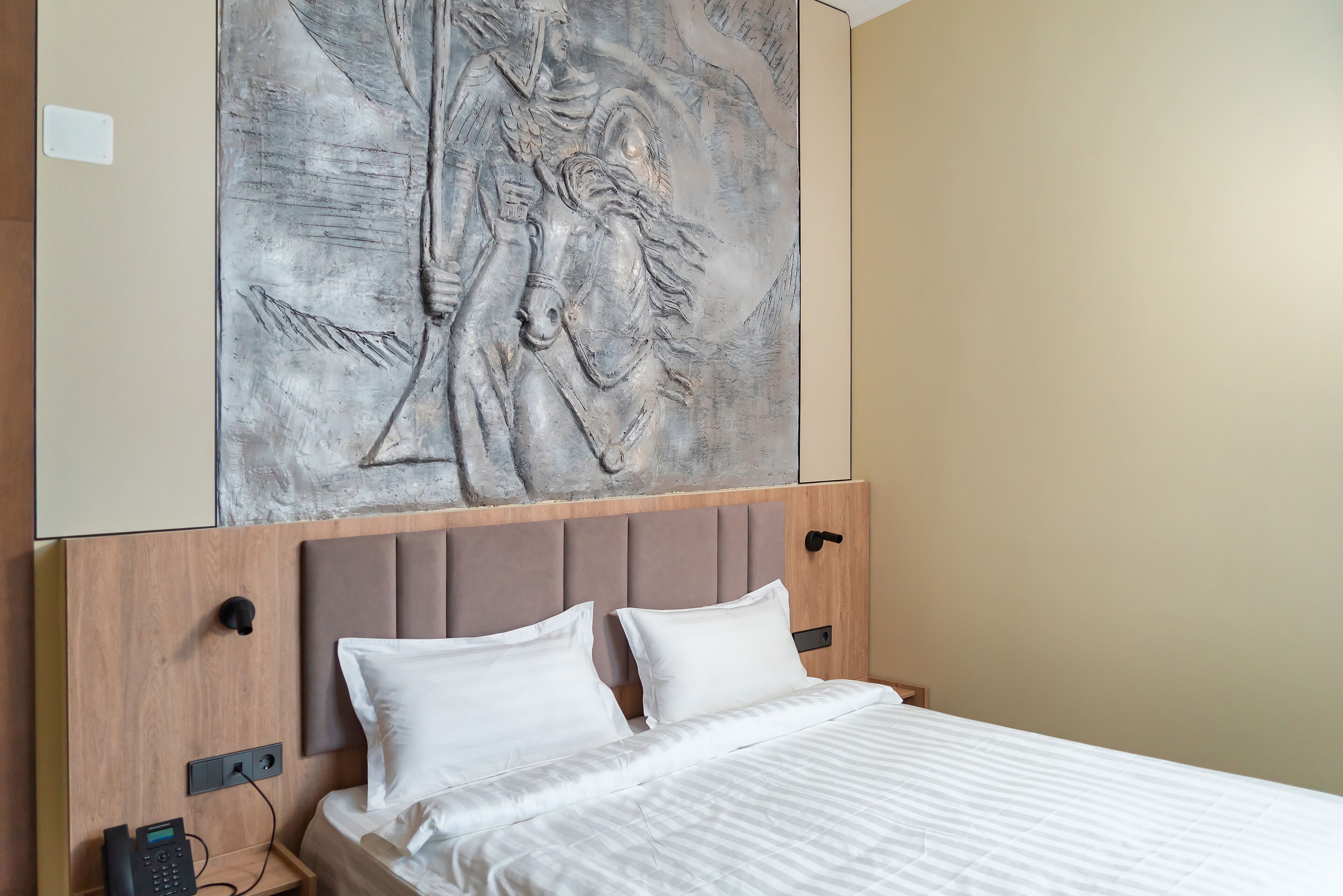 Una habitación de hotel con una cama, un cabecero con un teléfono y una obra de arte colgada en la pared.