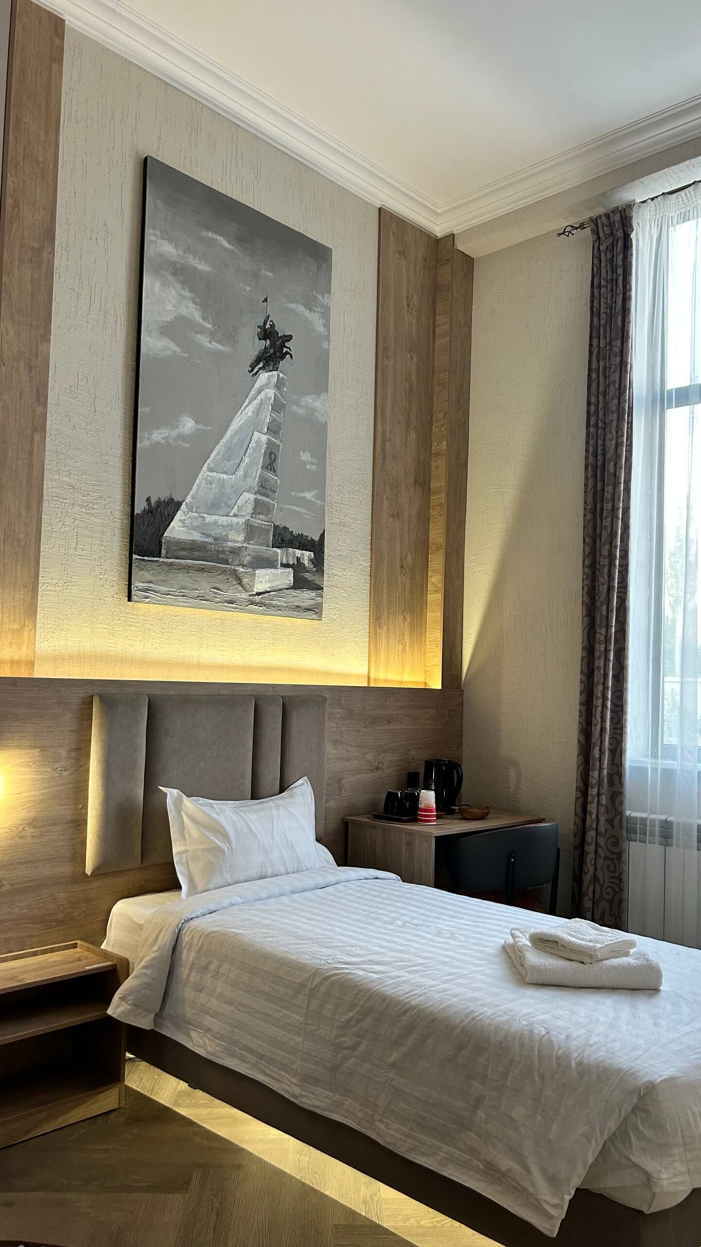 Una habitación de hotel con cama, mesita de noche, cafetera, un cuadro sobre la cama y una ventana con cortinas.