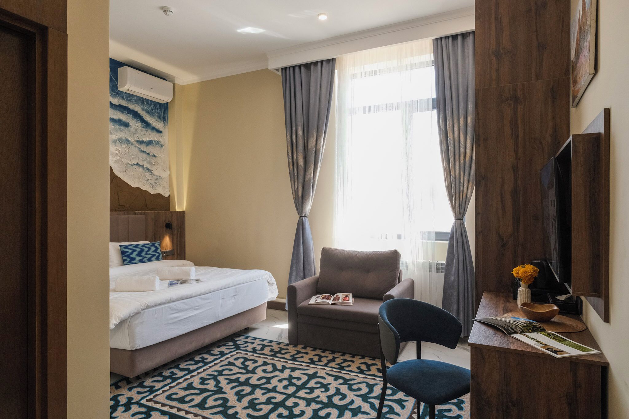 A hotel room with a bed, a sofa, a chair, a desk with a vase of flowers, and a large window with curtains.