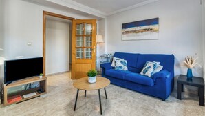Un salón con un sofá azul, un televisor sobre un mueble de madera, una mesa de centro redonda con una planta, y una puerta que da a otra habitación.
