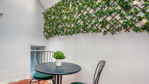 Un pequeño espacio al aire libre con una mesa redonda y dos sillas, una planta en maceta y una planta trepadora verde en una pared blanca.