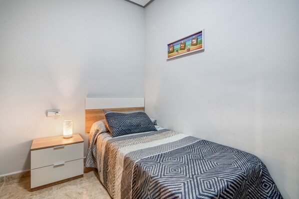 Un dormitorio con cama, mesita de noche con lámpara, un cuadro en la pared y una cómoda pequeña.