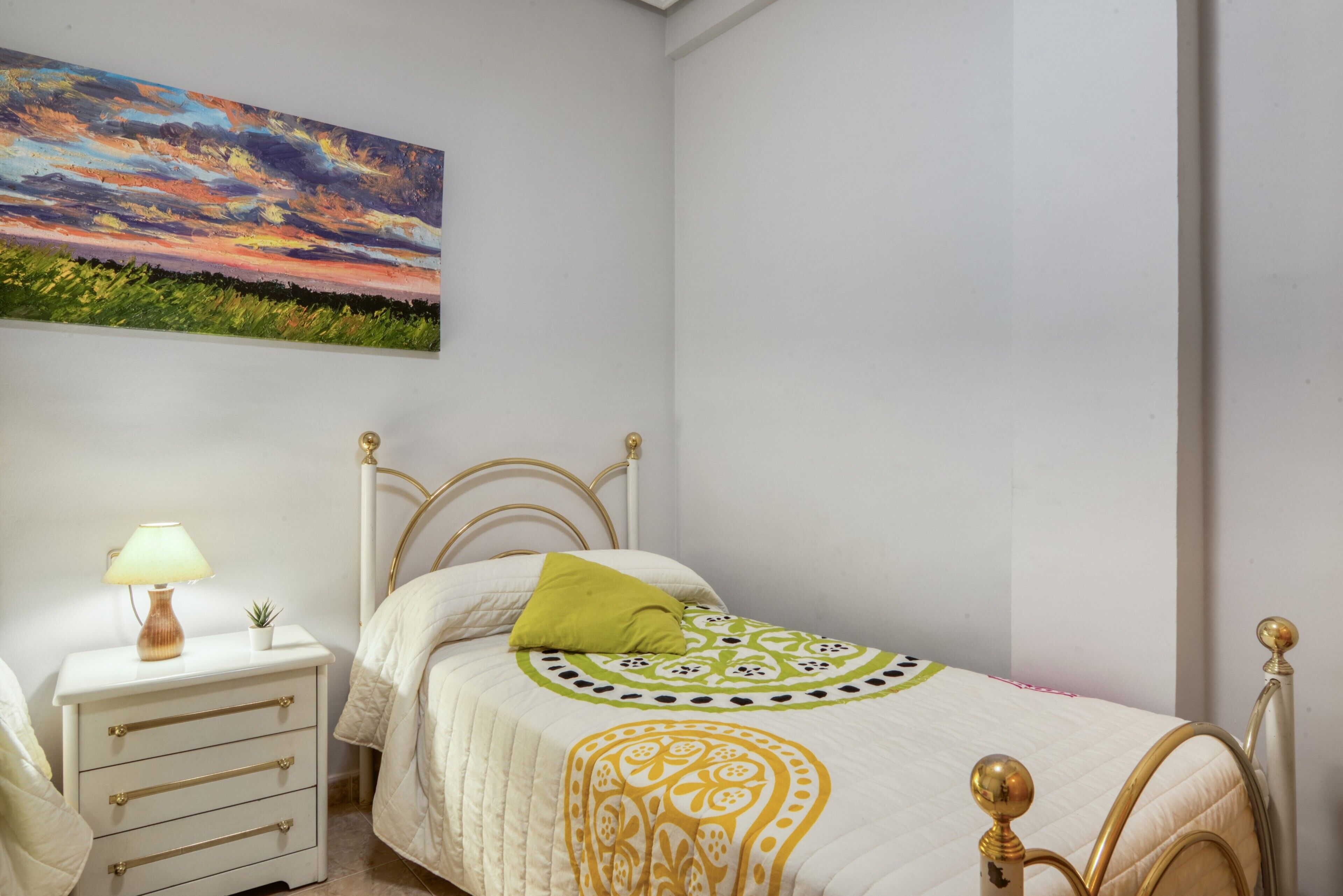 Un dormitorio con cama, mesita de noche, lámpara y un cuadro en la pared.