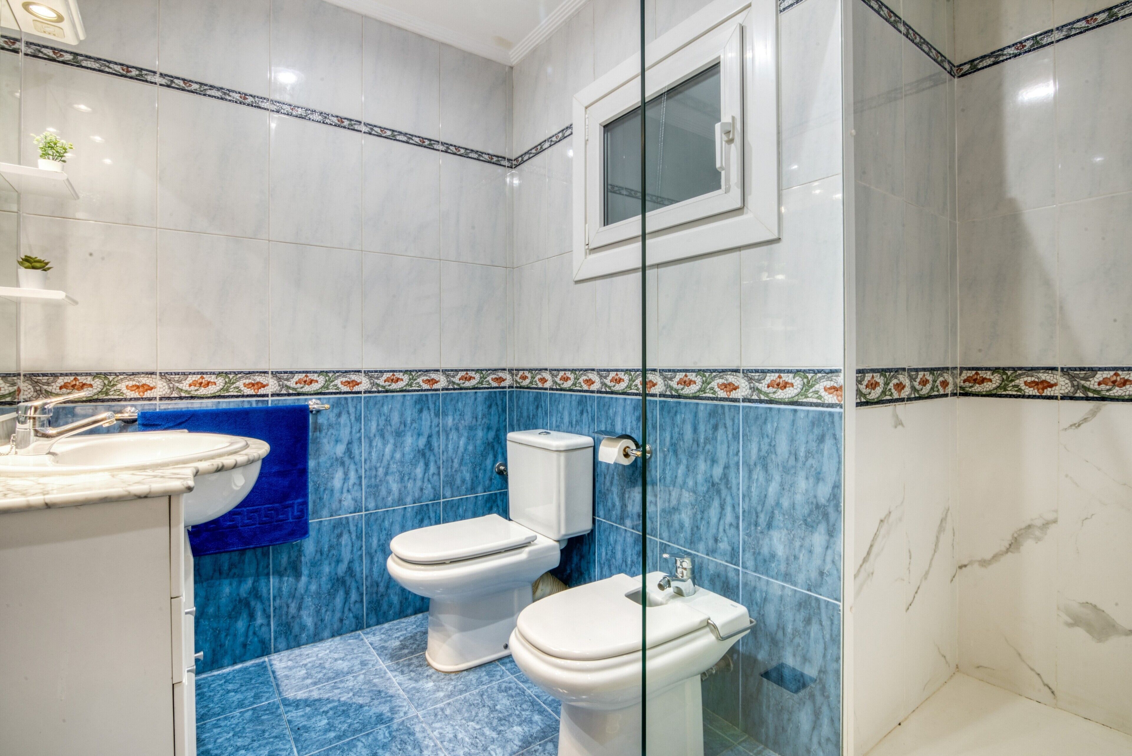 Un bany amb vàter, bidet i rentapel, rajoles blaves i blanques, i una finestra.