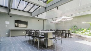 Un área de comedor exterior moderna con techo alto y tragaluces, que incluye una mesa grande y sillas, y un televisor montado en la pared.