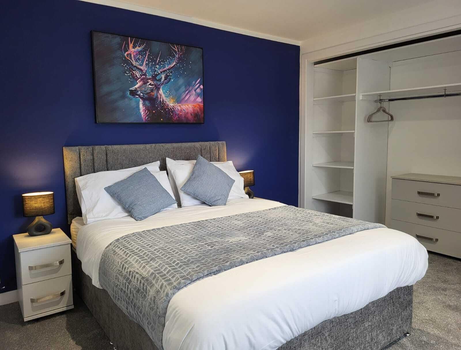 A bedroom with a large bed, a nightstand with a lamp, a wardrobe, and a painting on the wall.