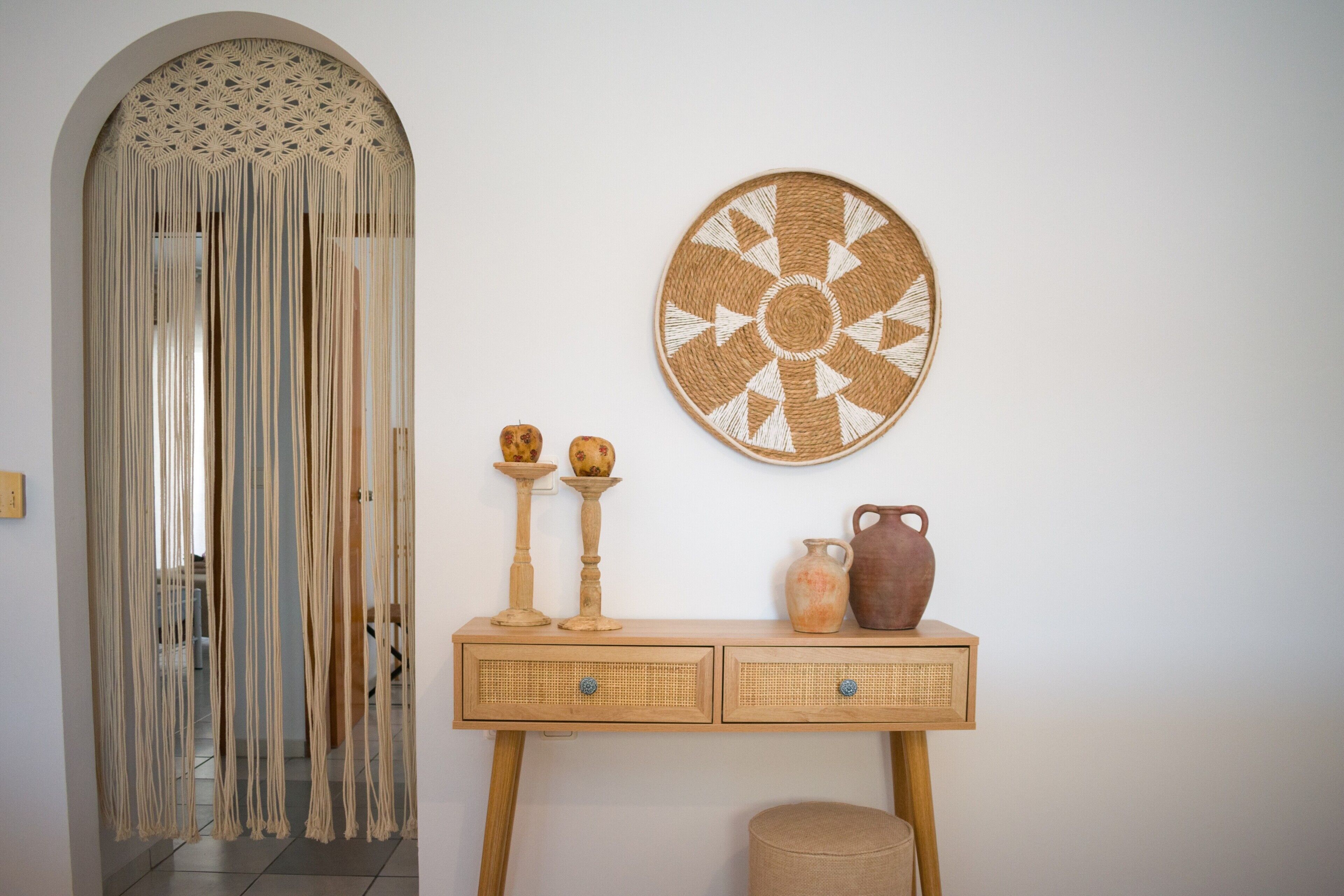 A room with a macramé wall hanging, a woven wall art piece, a wooden console table with drawers, and a small stool.