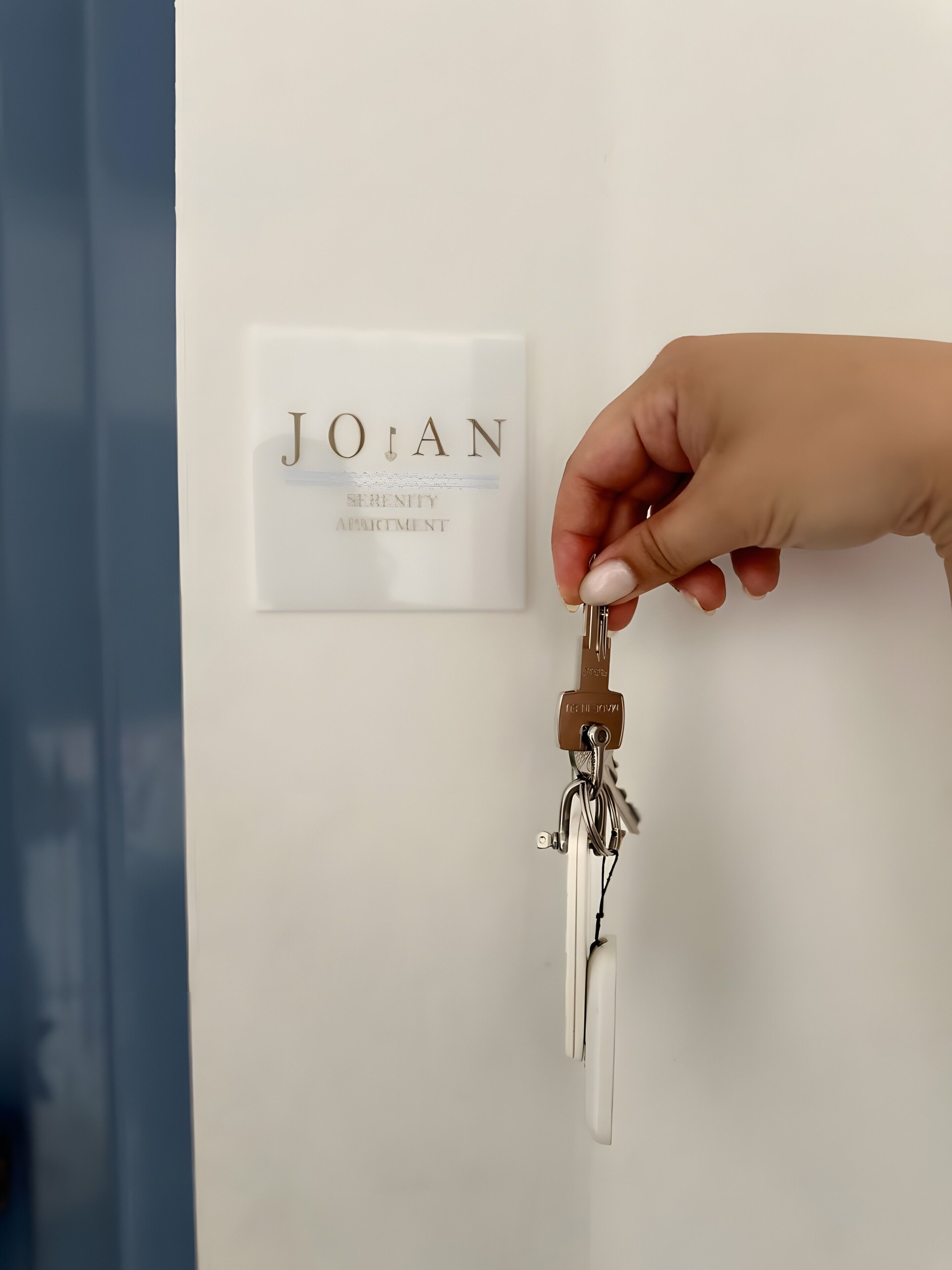 Una mà sosté una clau amb una placa que diu 'JOAN' i 'APARTAMENT SERENITY' a una porta blanca.