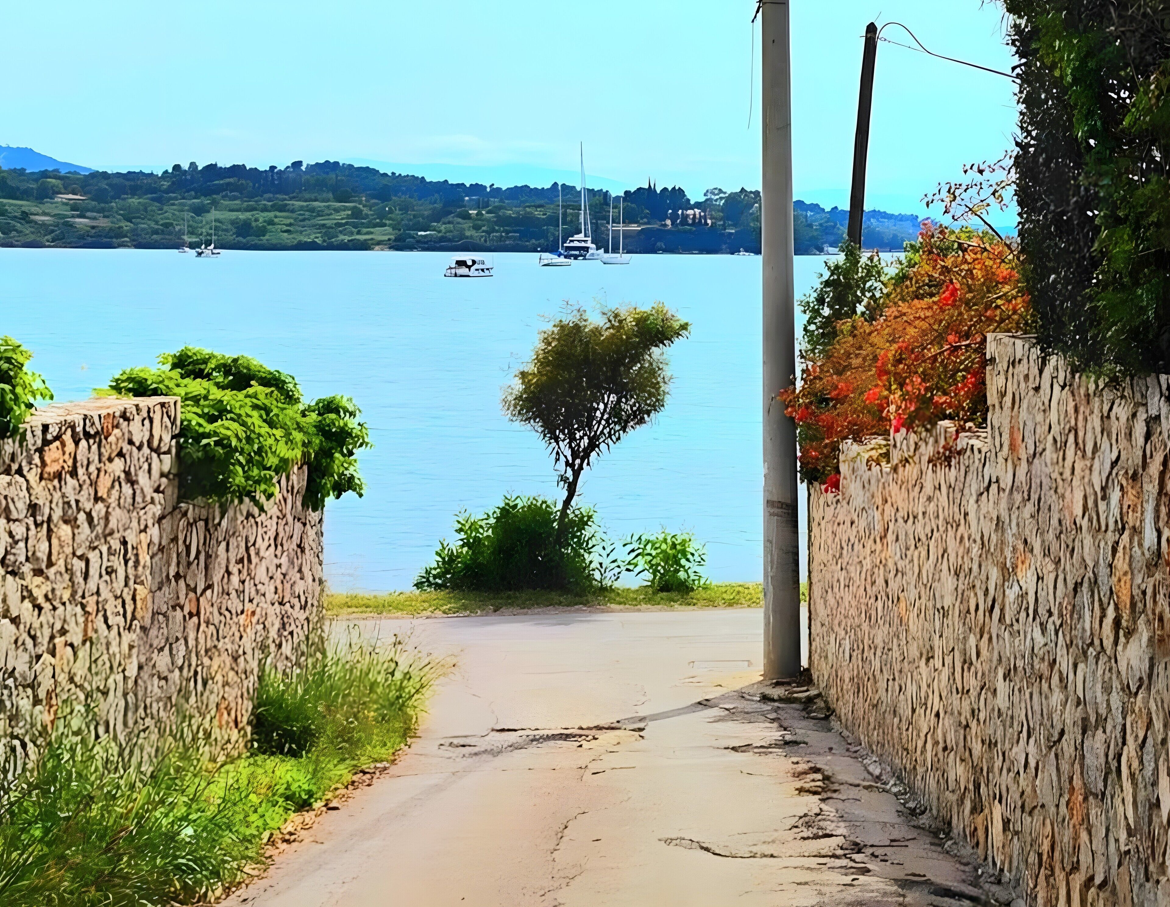 A pathway leading to a body of water with boats, surrounded by stone walls and greenery.