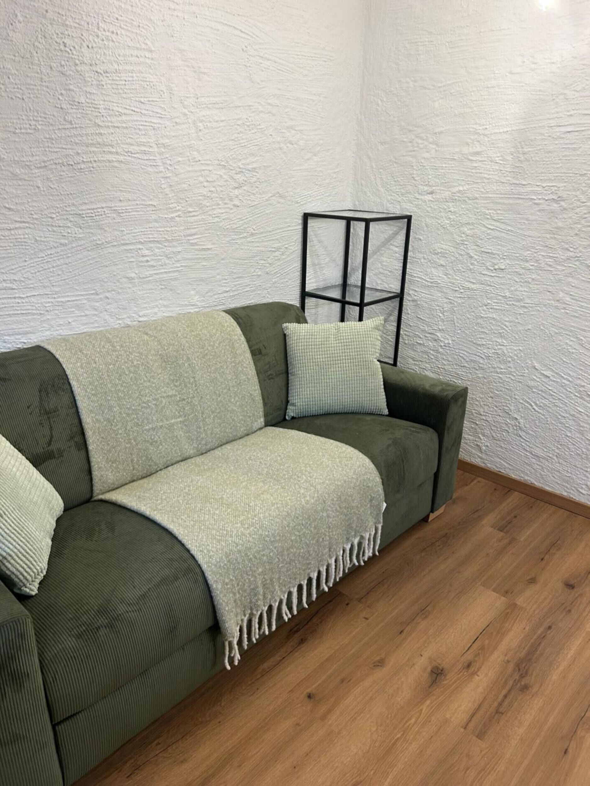 A green sofa with a throw blanket and pillows, a black metal side table, and a wooden floor.