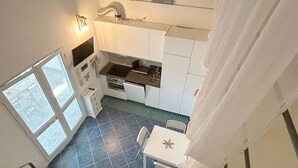 Une cuisine avec des armoires blanches, une cuisinière et un évier. Il y a une fenêtre sur la gauche et une table avec des chaises au centre.