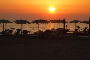 A beach with rows of sun loungers and umbrellas, a boat, and the sun setting over the water. - Apartment Rifugio in Pineta near the sea (Marina di Grosseto)