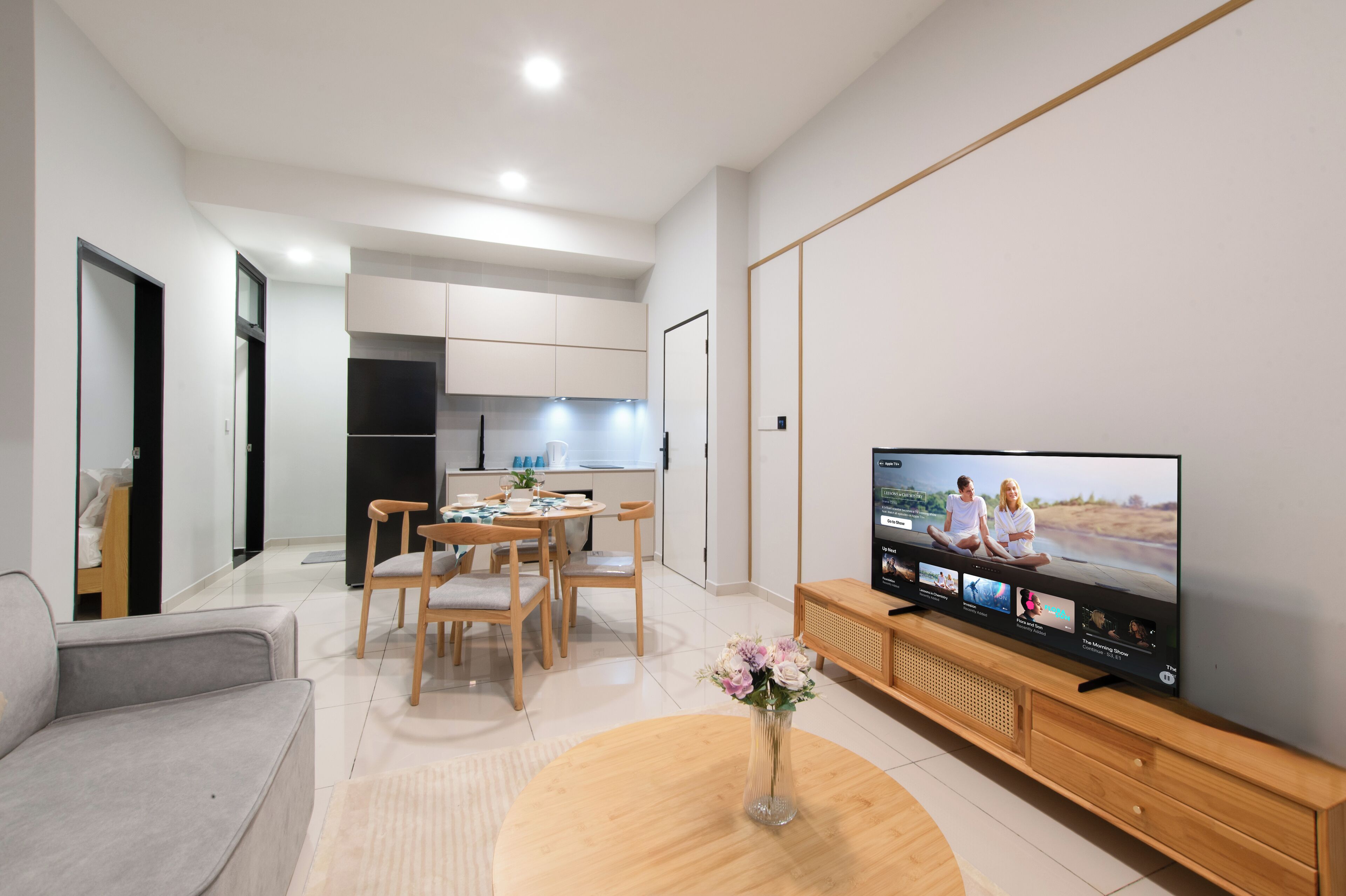 A modern living room with a flat-screen TV, a dining area with wooden chairs, and a kitchen in the background.