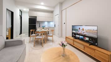 A modern living room with a flat-screen TV, a dining area with wooden chairs, and a kitchen in the background.