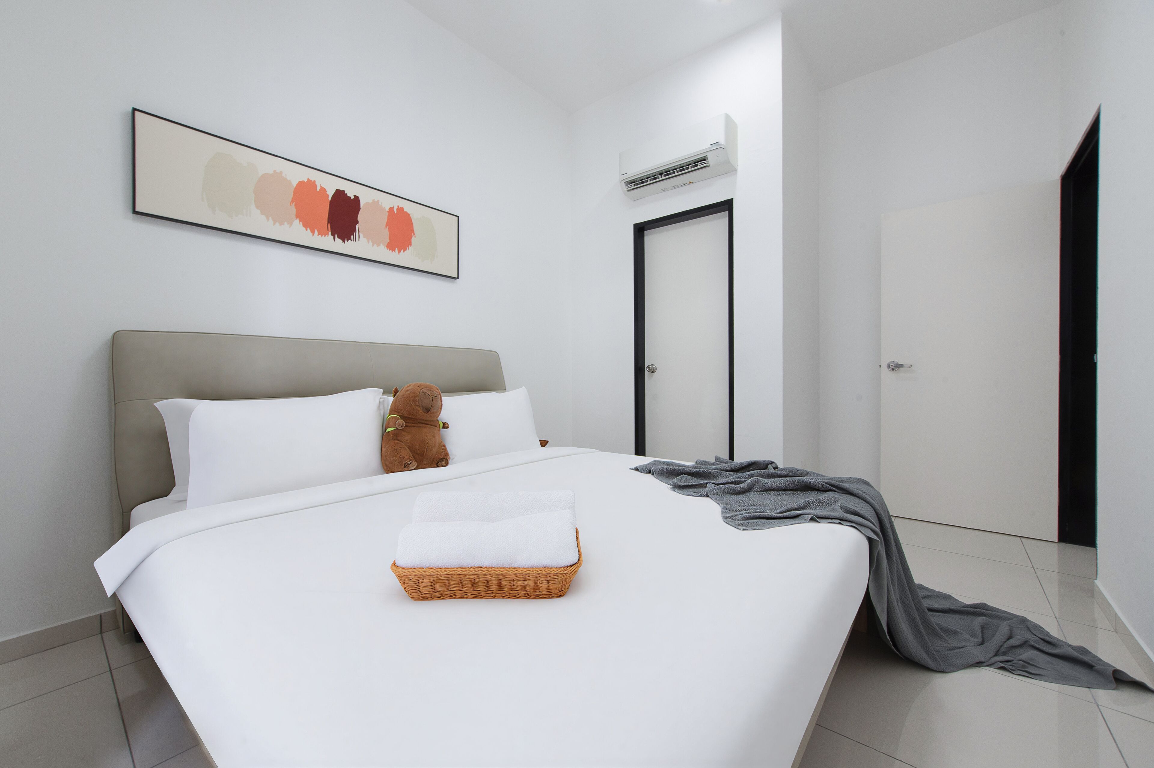 Habitación de hotel moderna con una cama grande, un osito de peluche, una obra de arte enmarcada, un equipo de aire acondicionado y una puerta.