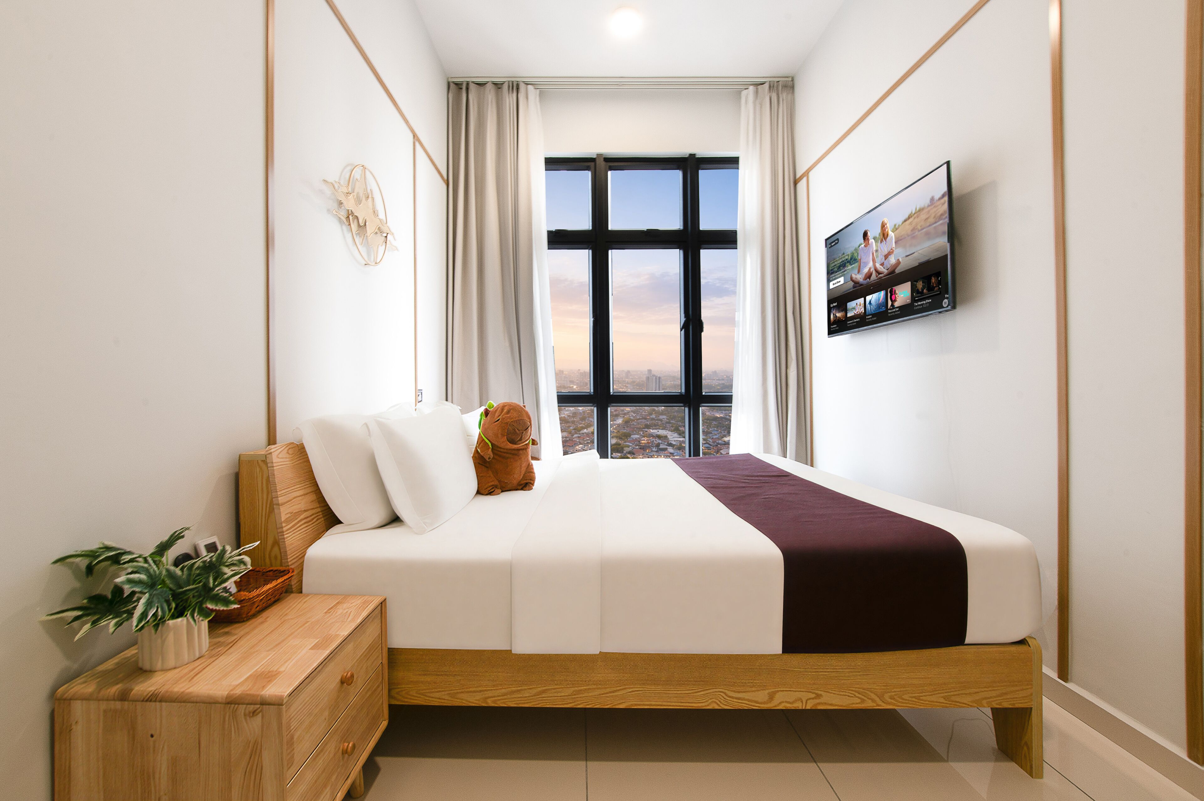 A modern bedroom with a large window, a bed with white bedding, a wooden nightstand, and a television on the wall.